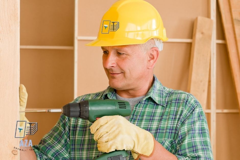 A drywall repair company worker in a yellow hard hat and green plaid shirt holds a power drill while standing next to stacked drywall sheets in a residential construction setting near W Peggy Dr, and N Lepa Dr, San Tan Valley, AZ.