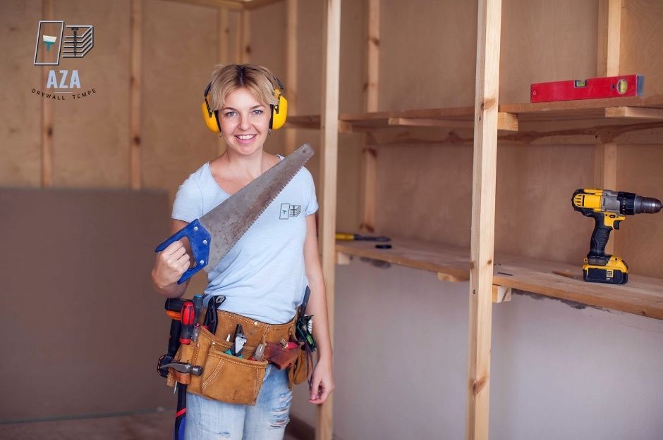 A drywall repair handyman in a white t-shirt and tool belt holds a hand saw inside an unfinished room with exposed wooden framing. A red level rests on a shelf, showing tools ready for residential construction near W Rose Canyon Cir and W Meridian Dr, Anthem, AZ.