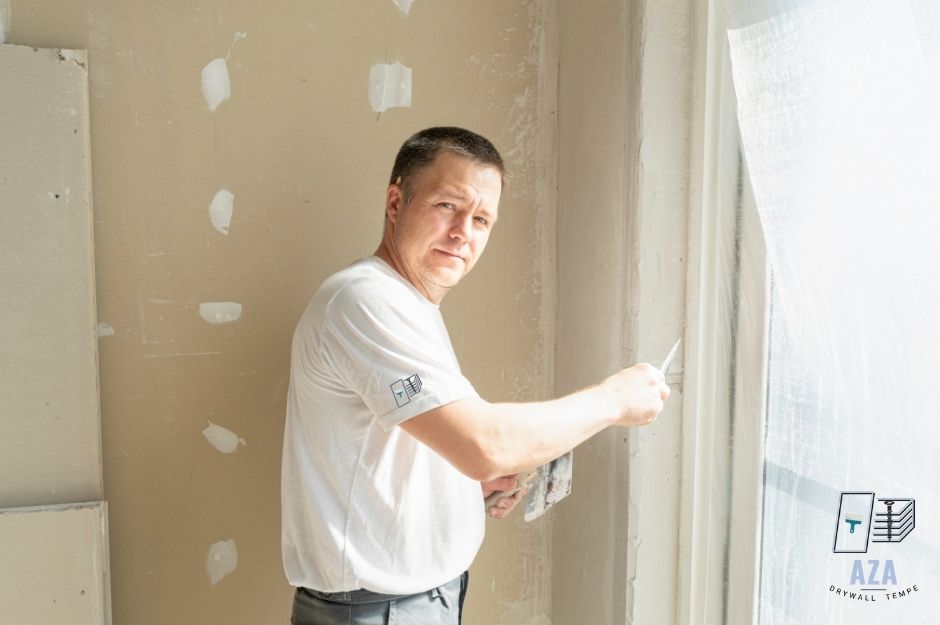 A drywall repair handyman at a commercial construction site holds a putty knife while working on wall patches in an unfinished room with exposed drywall and repair spots near E Calle San Angelo, and Cll Moctezuma, Guadalupe, AZ.