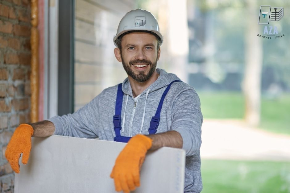 A drywall repair handyman in a gray hoodie and white hard hat smiles, wearing orange work gloves and leaning against white drywall sheets at a construction site. He wears blue overalls in a residential setting near S 43rd Ln, and W Piedmont Rd, Laveen, AZ.