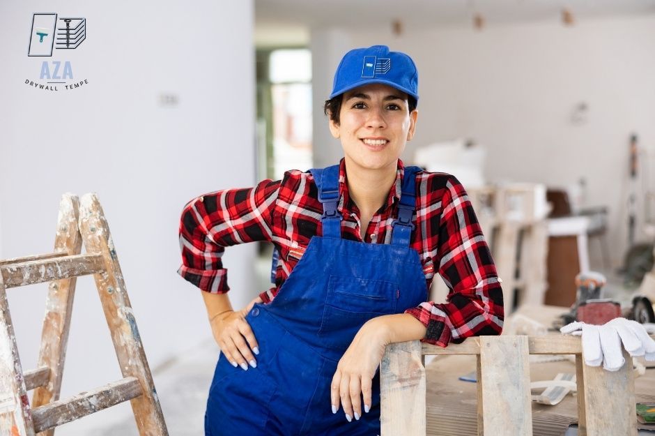 A drywall repair specialist wearing a blue cap, plaid shirt, and overalls poses confidently in a residential renovation space. She leans casually against a wooden ladder with construction materials and tools in the background near N 178th Ave, and W Northern Ave, Waddell, AZ.