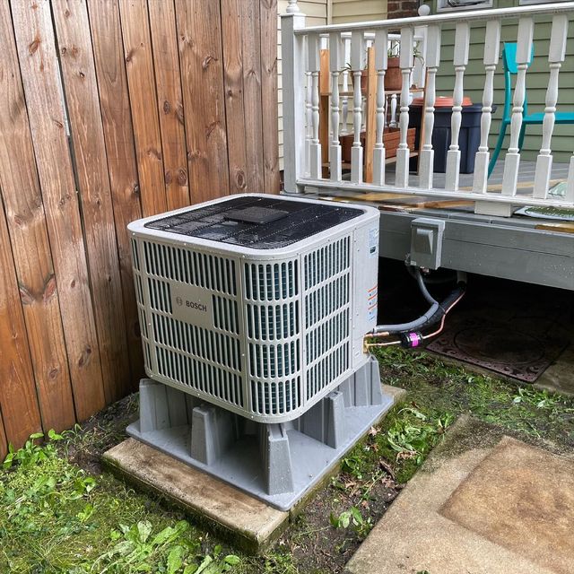 HVAC unit on a concrete pad next to a wooden fence and deck. Gray, beige, and green.