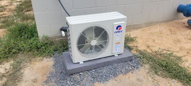 An air conditioning unit on a concrete base, set on gravel and dirt, next to a building.