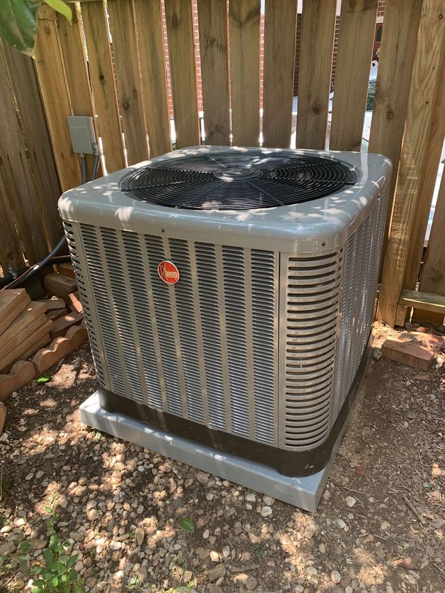 Gray Rheem air conditioning unit outside by a wooden fence.