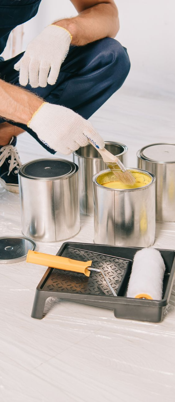 A person wearing gloves stirs yellow paint in a metal can, with a paint roller and tray placed on the floor nearby.