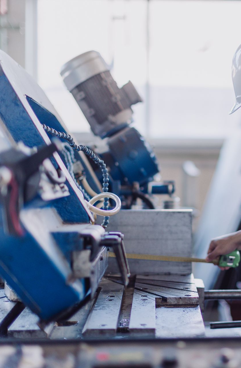 A worker uses a tape measure to check the dimensions of a metal piece on a blue industrial band saw.
