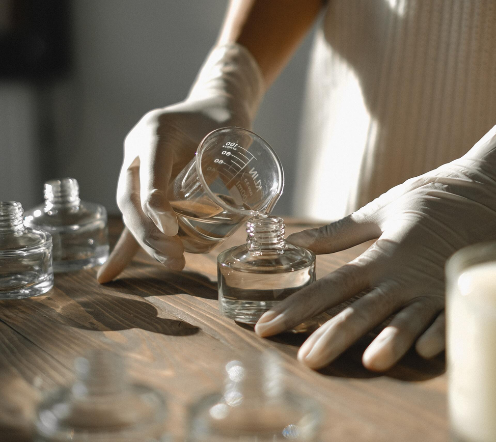 Hands in gloves pour clear liquid from a beaker into a small glass bottle on a wooden table.