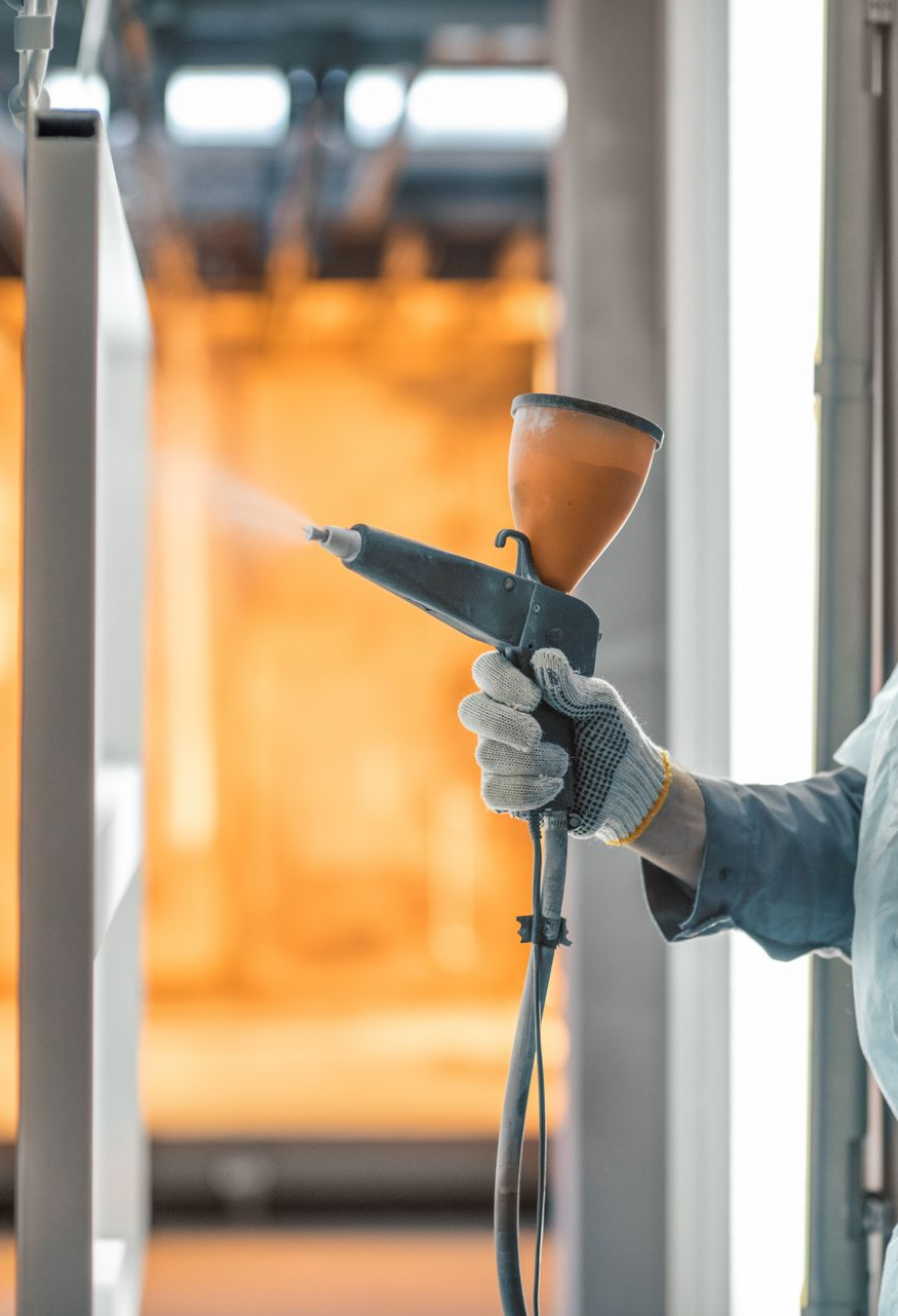 A person wearing a glove uses a handheld powder coating spray gun to apply finish to a metal surface in an industrial area.