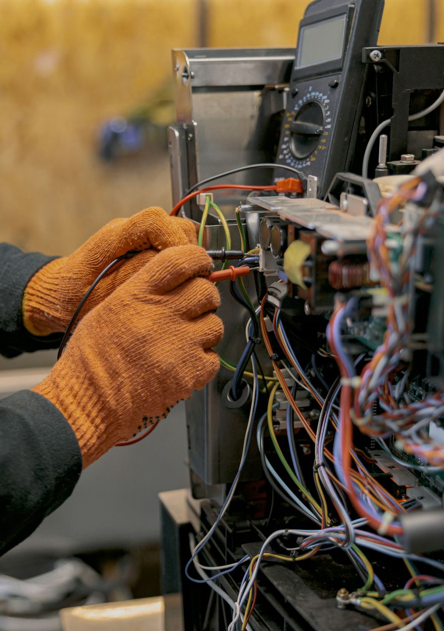 A person wearing orange work gloves uses a multimeter to test electrical components inside a device.