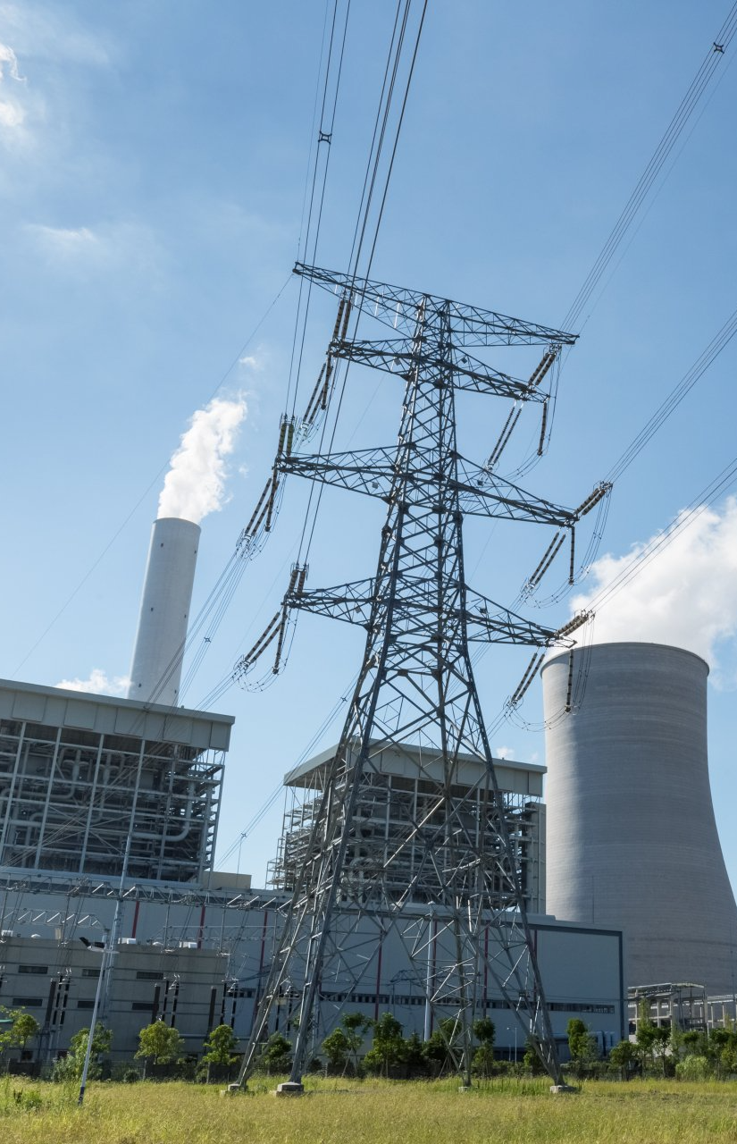 A metal electrical transmission tower stands before a power plant with a tall chimney and a large cooling tower.