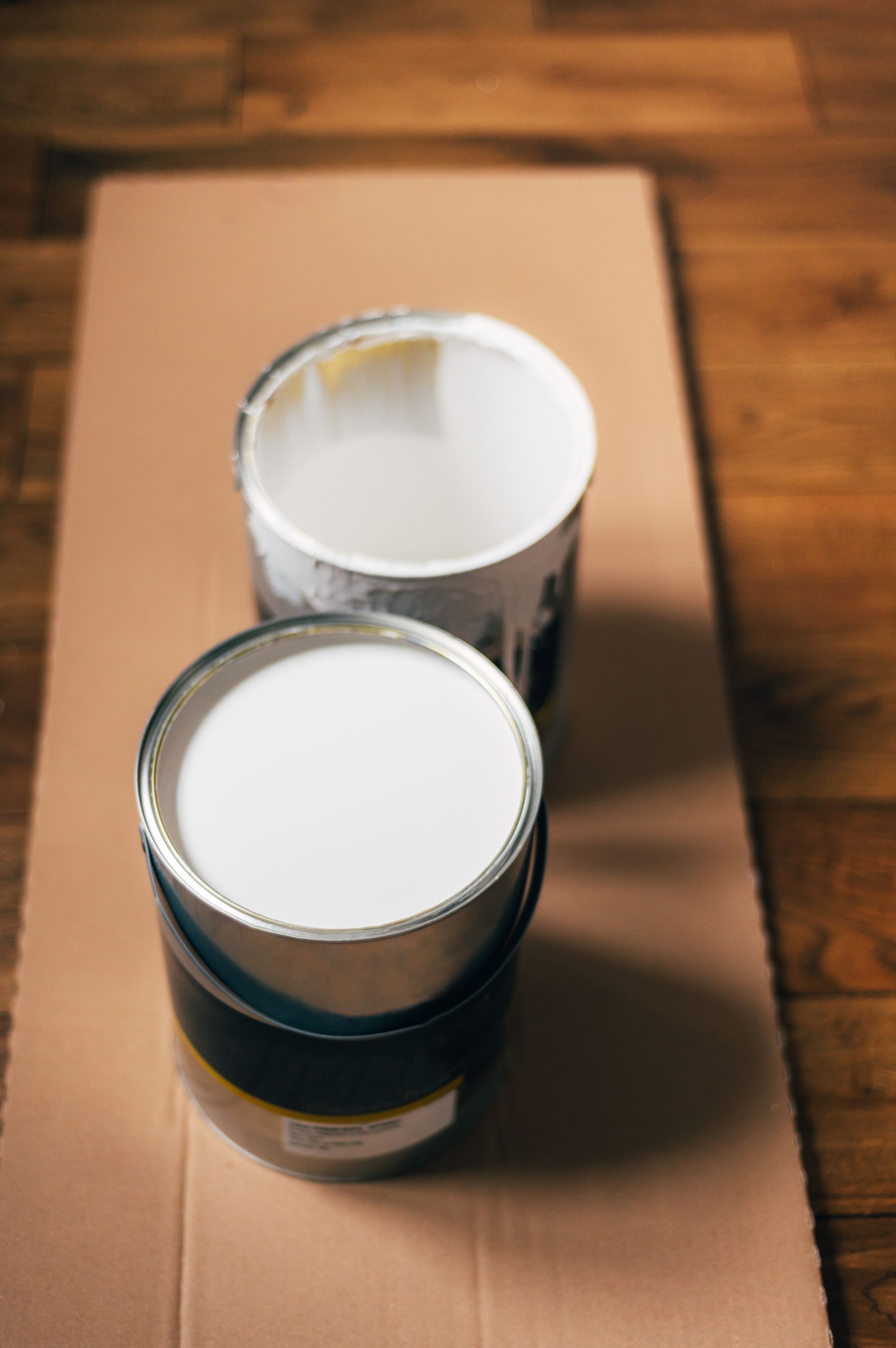 Two open paint cans, one full of white paint and one empty, sitting on cardboard on a wood floor.