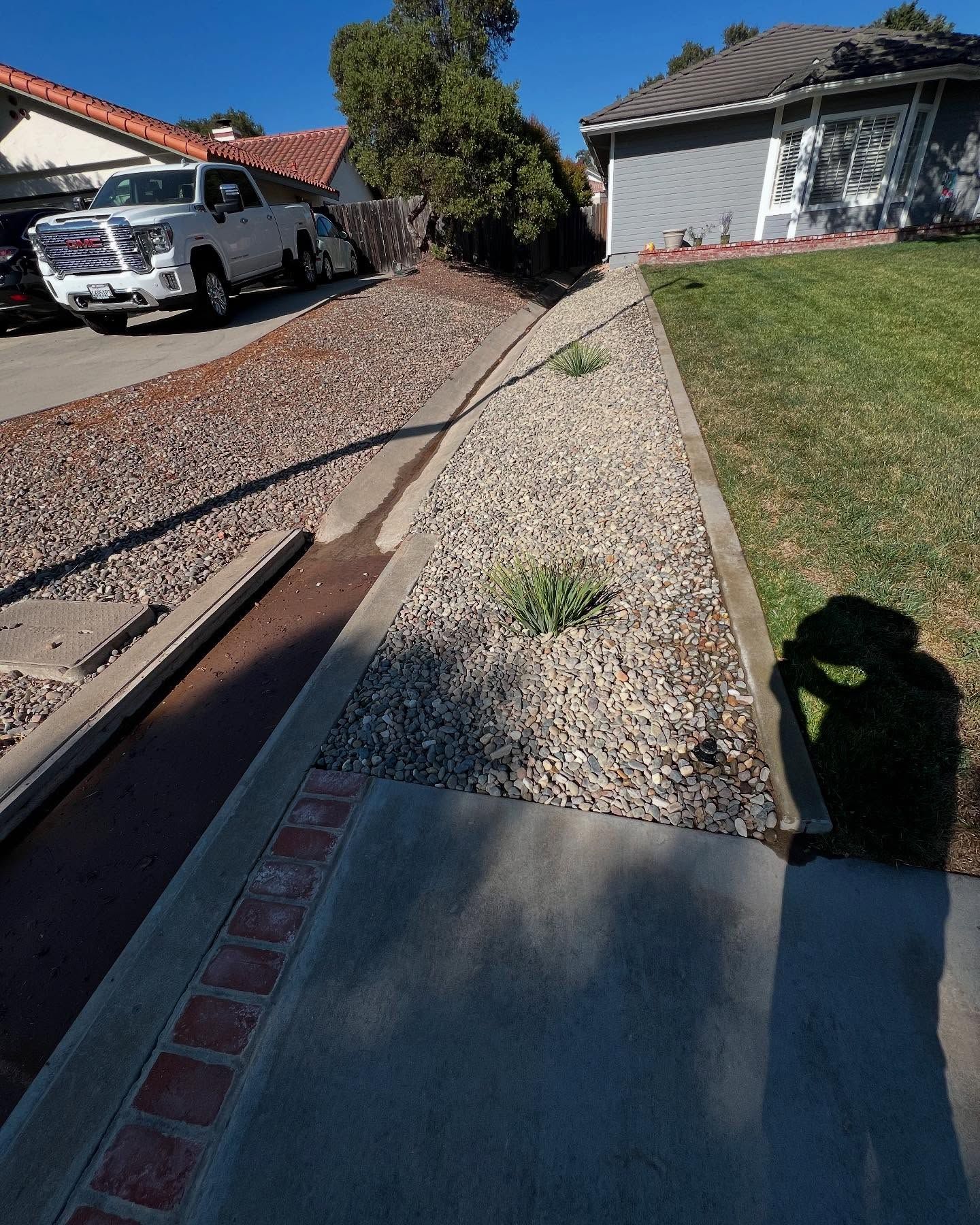 A driveway with a rock bed, sidewalk, grass, and a white truck. A shadow is cast on the grass.