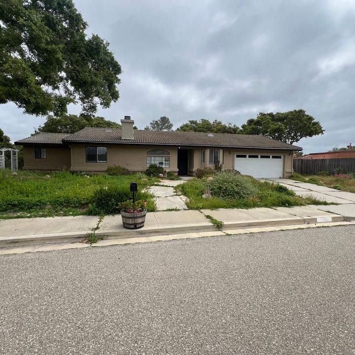 Ranch-style house with overgrown yard, cracked walkway, and cloudy sky.