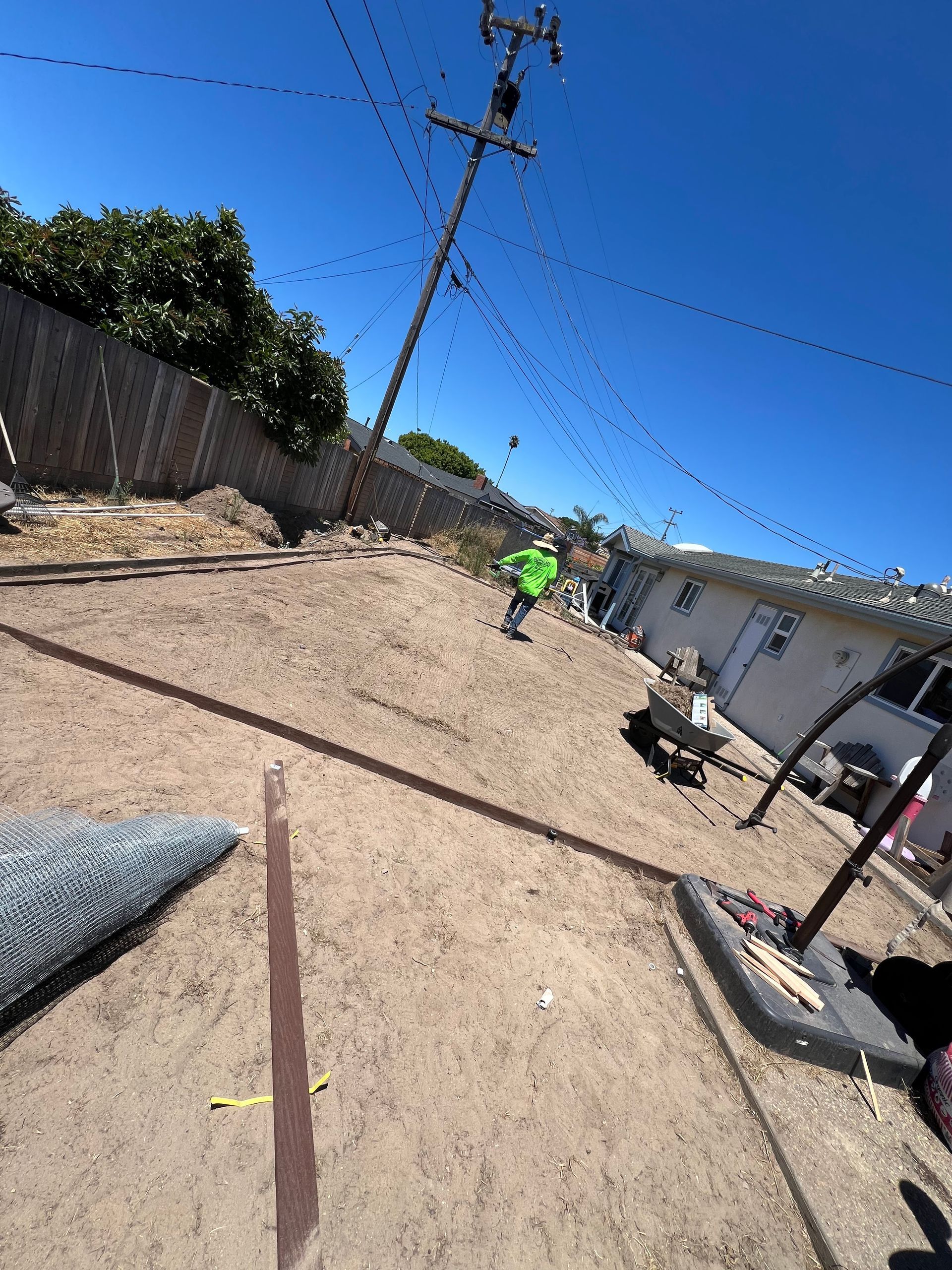 A person in green shirt working in a dirt backyard under a bright blue sky, with a power pole.