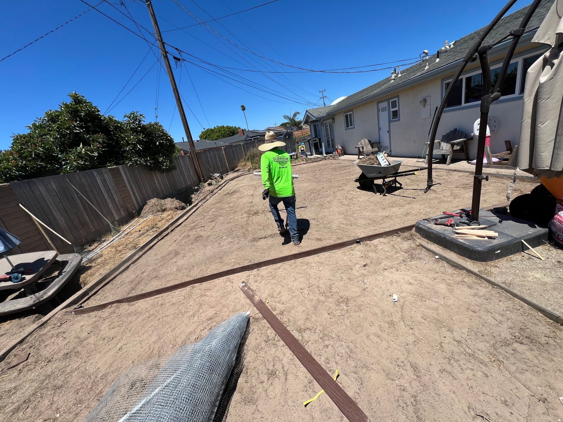 A person in a green jacket walks in a dirt backyard with a fence, power lines, and a building on a sunny day.