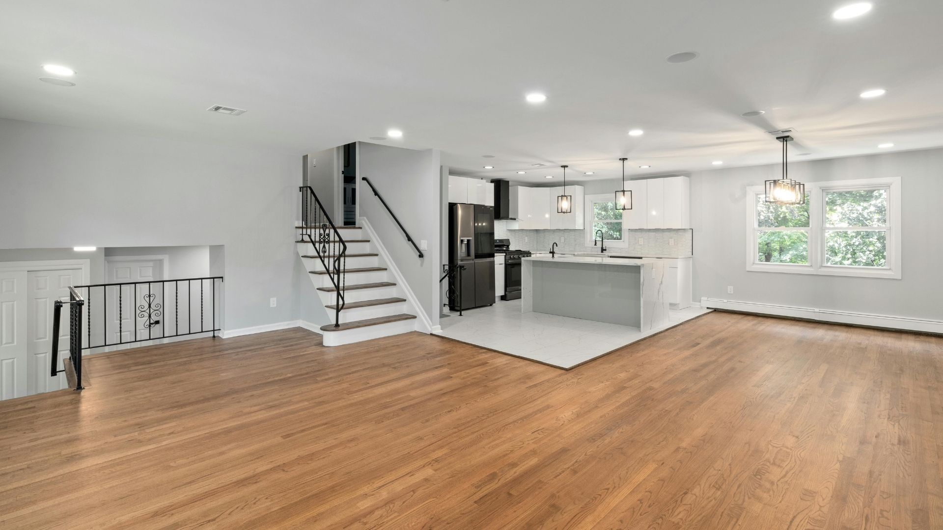 Modern open-plan living room with hardwood floors, white walls, staircase, and kitchen island with pendant lights.