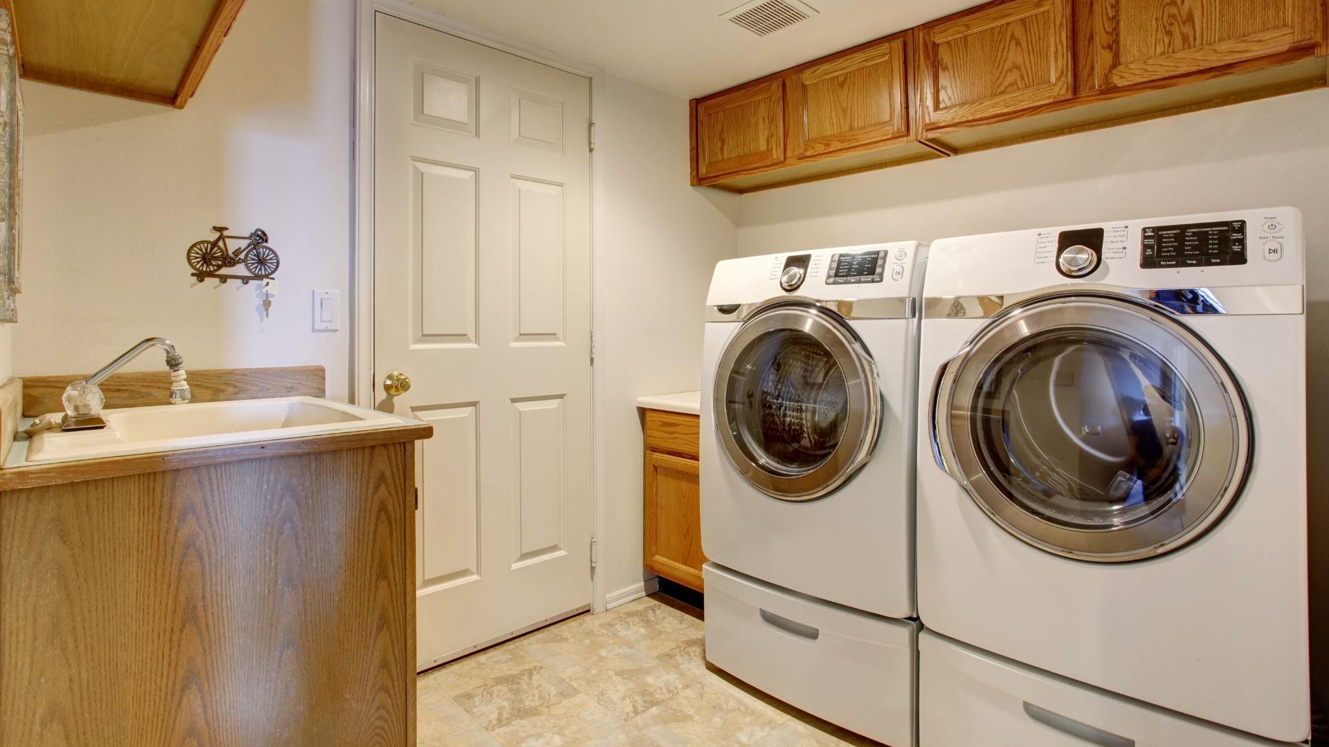 Laundry room with front-loading washer and dryer beside a utility sink and wooden cabinets.