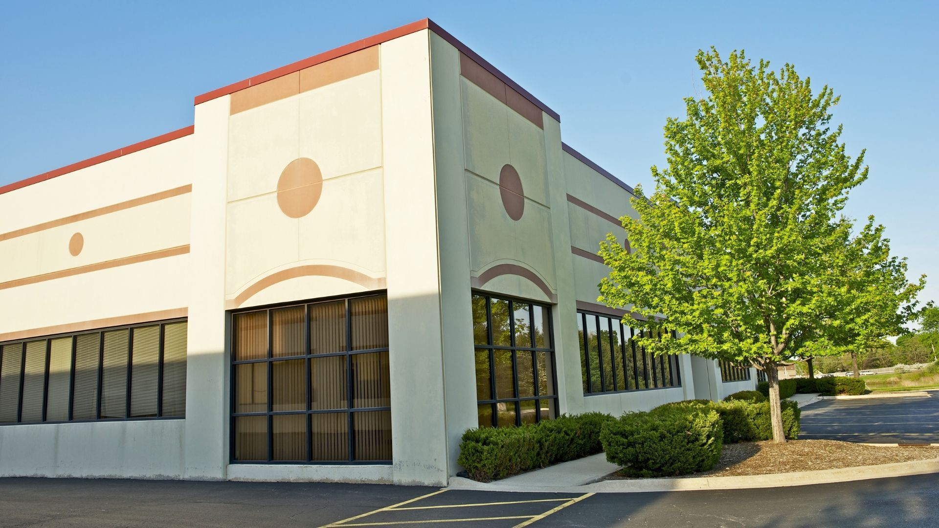 Corner of a beige commercial building with dark windows and a green tree beside a parking lot