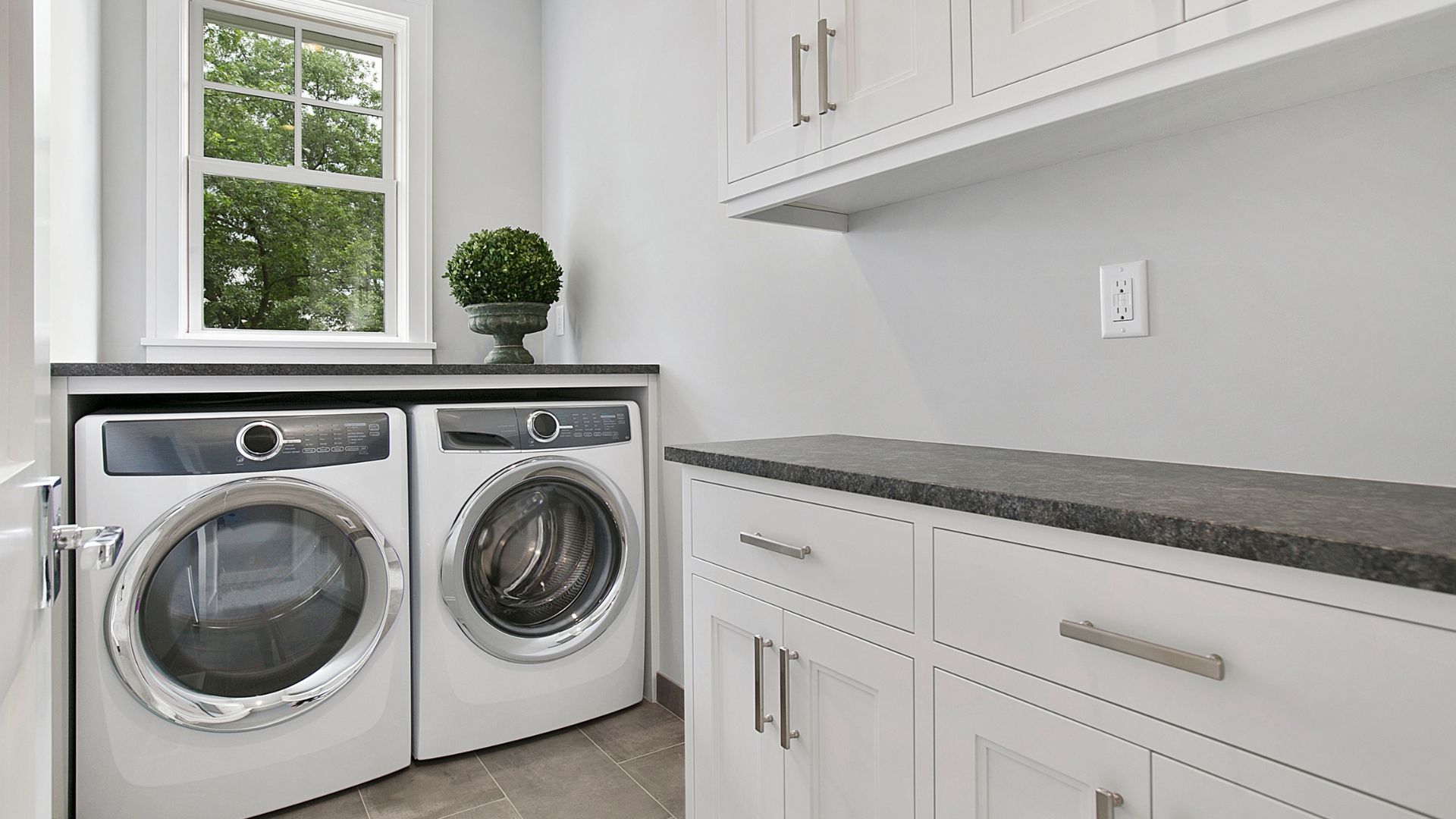 Laundry room with front-loading washer and dryer beside white cabinets and a countertop by a window.