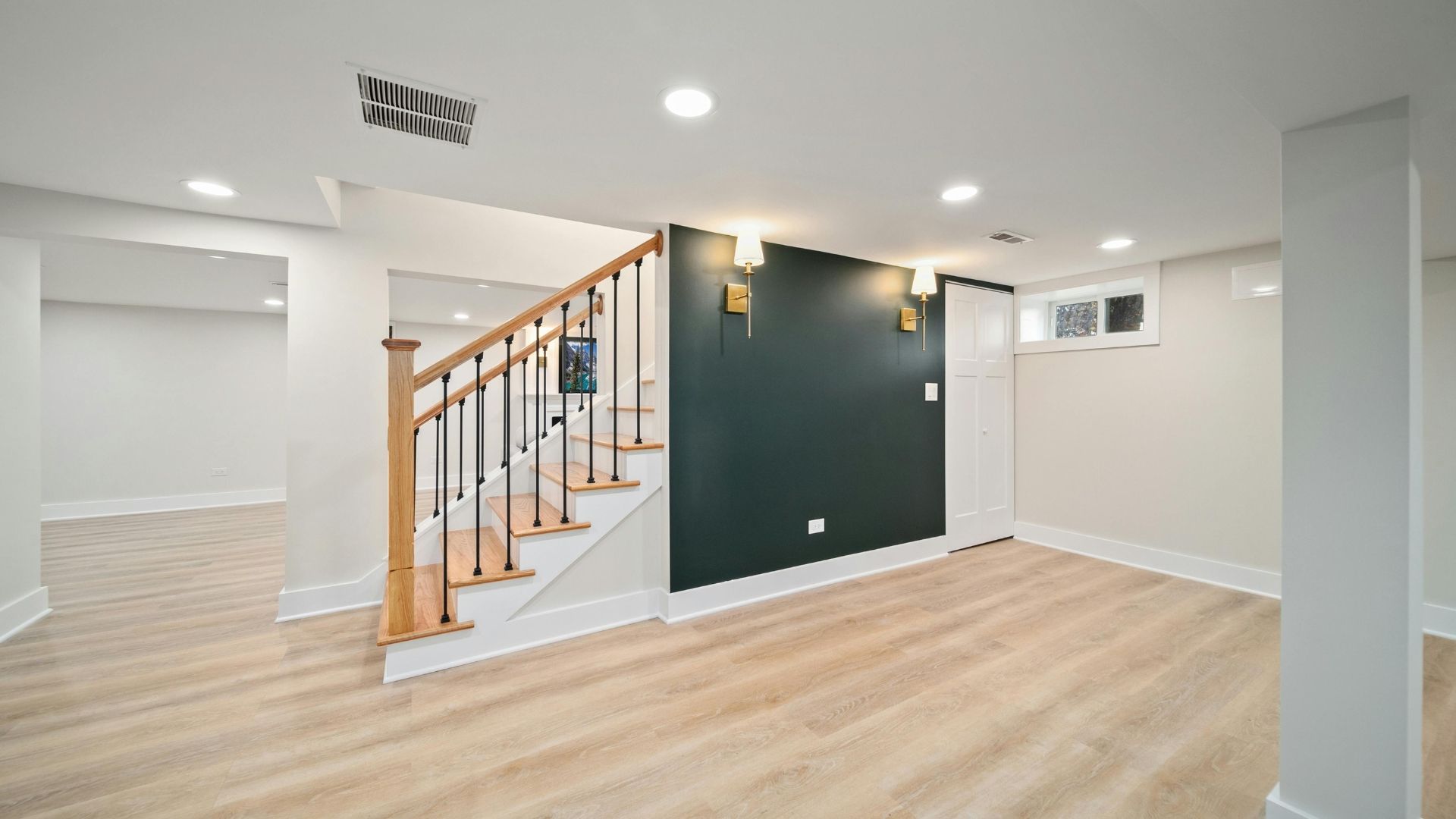 Bright empty basement room with light wood floors, white walls, and a staircase with wooden railing.