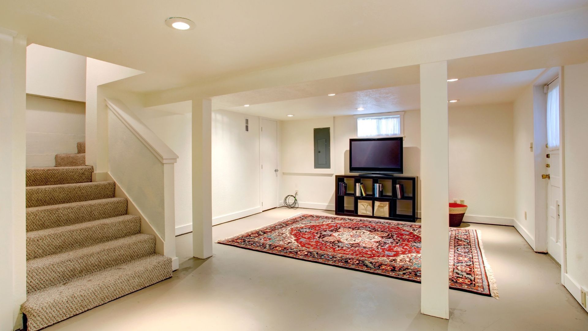 Finished basement with carpeted stairs, white columns, and a red patterned rug by a TV on a stand.