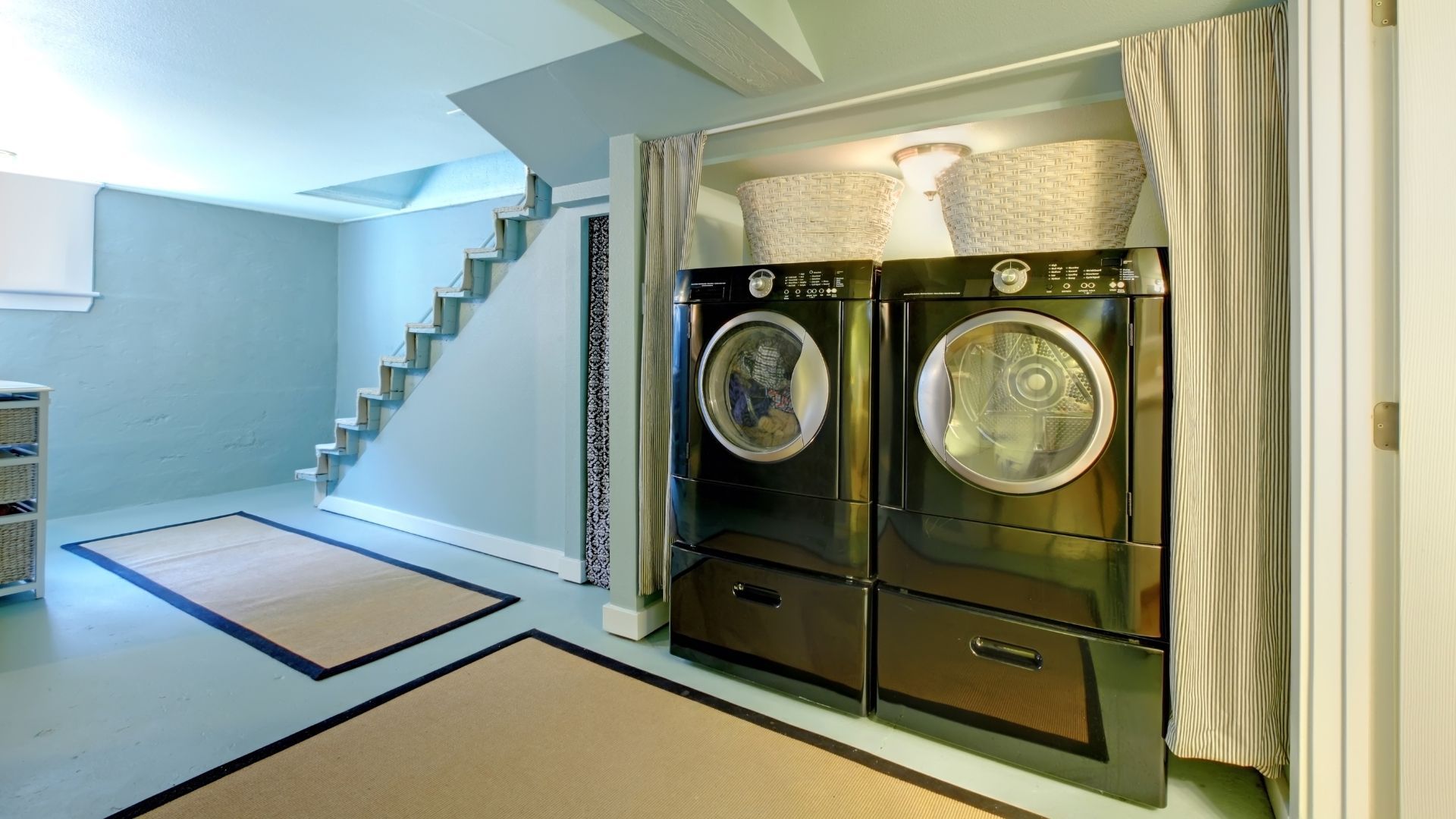 Laundry room with two black front-loading washers beside a staircase and curtained window.