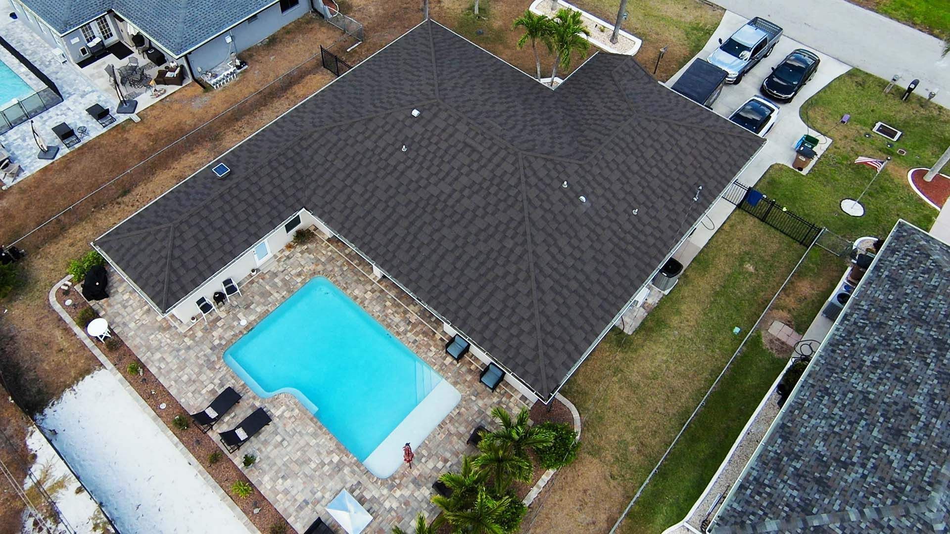 Aerial view of a house with a pool and dark roof, surrounded by a yard, with cars parked in the driveway.