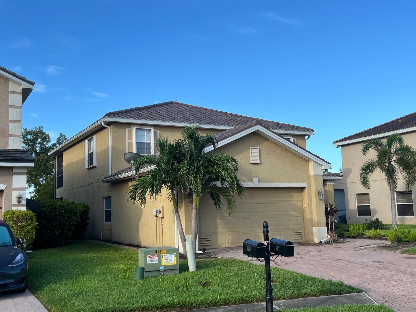 Tan two-story house with brown roof, mailbox in front, and palm trees under a clear blue sky.
