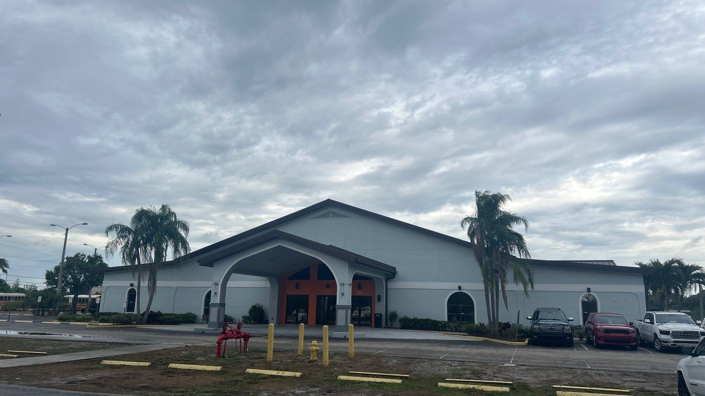 Exterior view of a light gray building with a dark roof and orange entrance under a cloudy sky. Cars are parked in front.