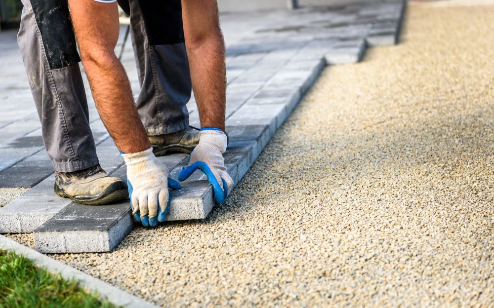 Person placing grey paving stone on a bed of gravel outdoors — Rhyno’s Concreting In Old Bar, NSW
