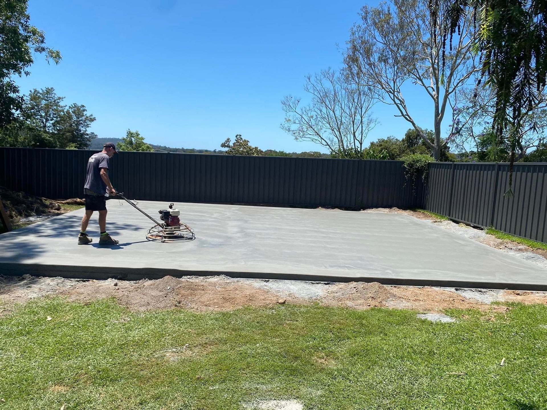 Man Smoothing Concrete with A Power Trowel, Preparing a Patio. Black Fence in The Background, Blue Sky — Rhyno’s Concreting In Old Bar, NSW