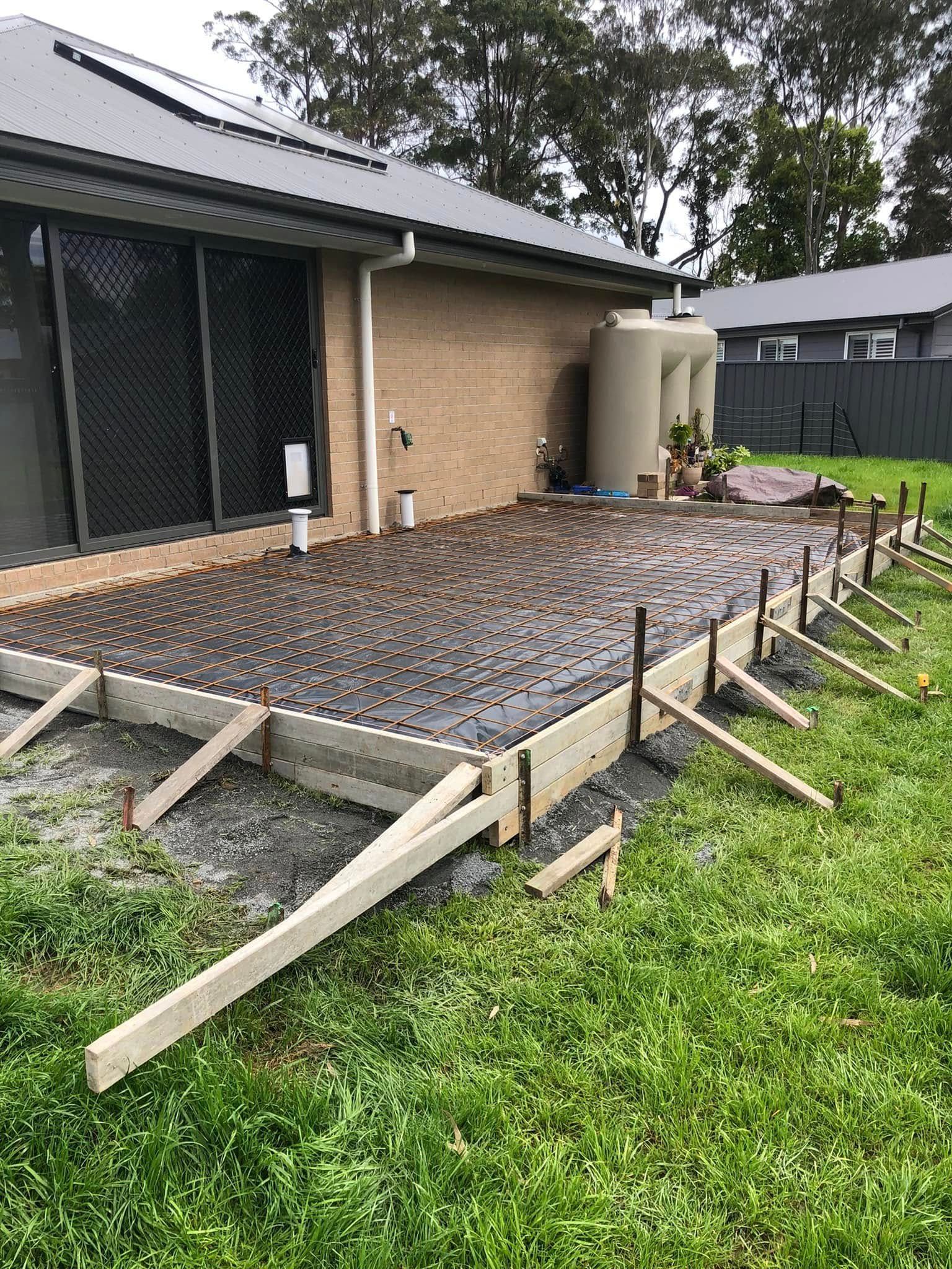 House side yard with a freshly poured concrete slab framed by wooden forms and a gravel surface.