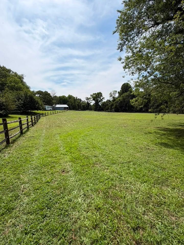 Grassy field beside a wooden fence under a partly cloudy sky, with trees and a small building in the distance