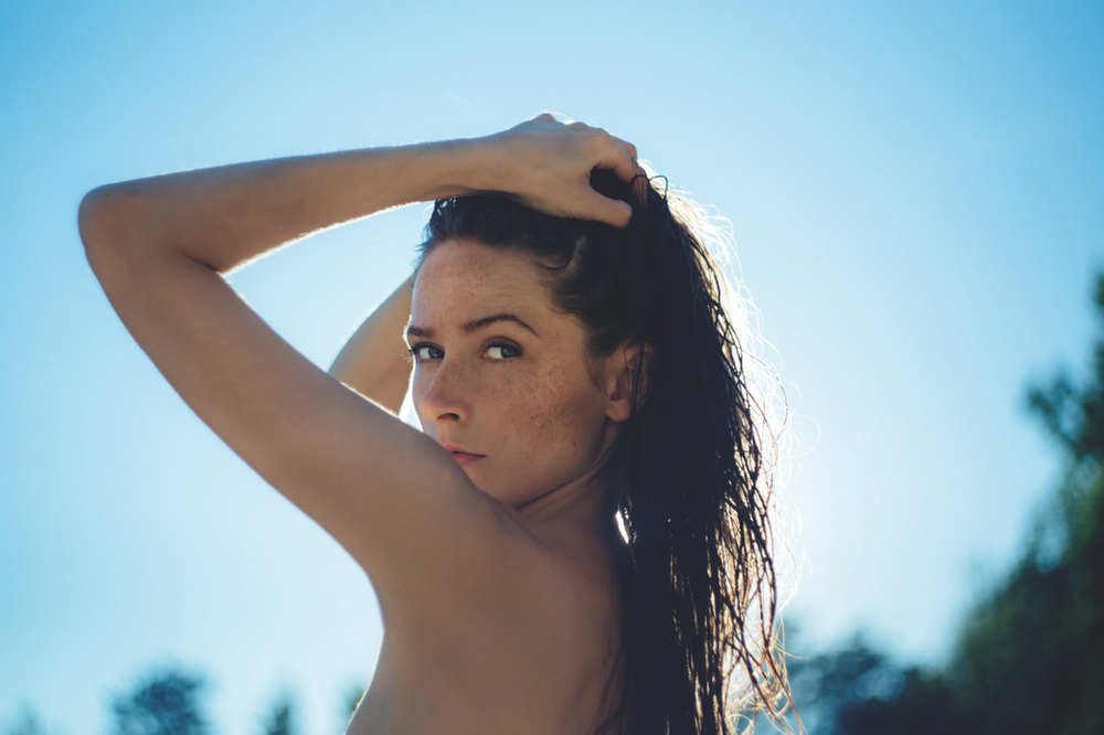 A woman without a shirt is holding her hair in front of a blue sky.