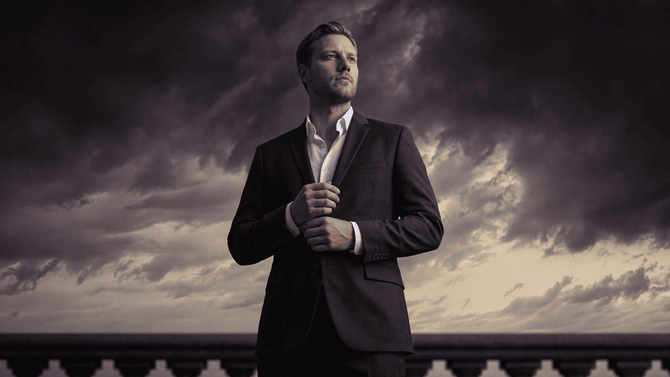 Man in a dark suit adjusting his jacket against a dramatic cloudy sky and railing