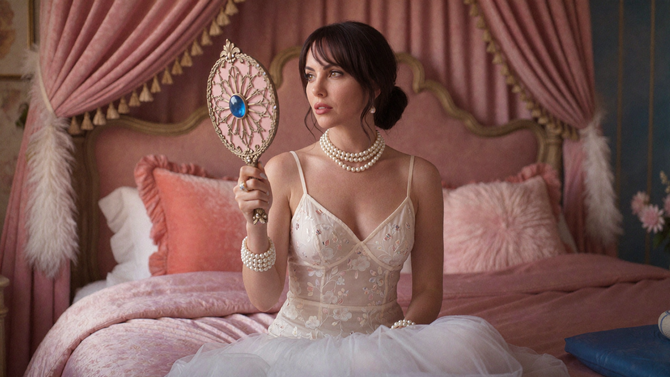 Woman in a pink bedroom holding a jeweled hand mirror, seated on a bed in a white dress.