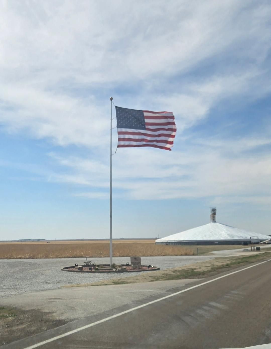 American flag on a tall pole near a roadside, with a white dome building and open field under a cloudy sky.