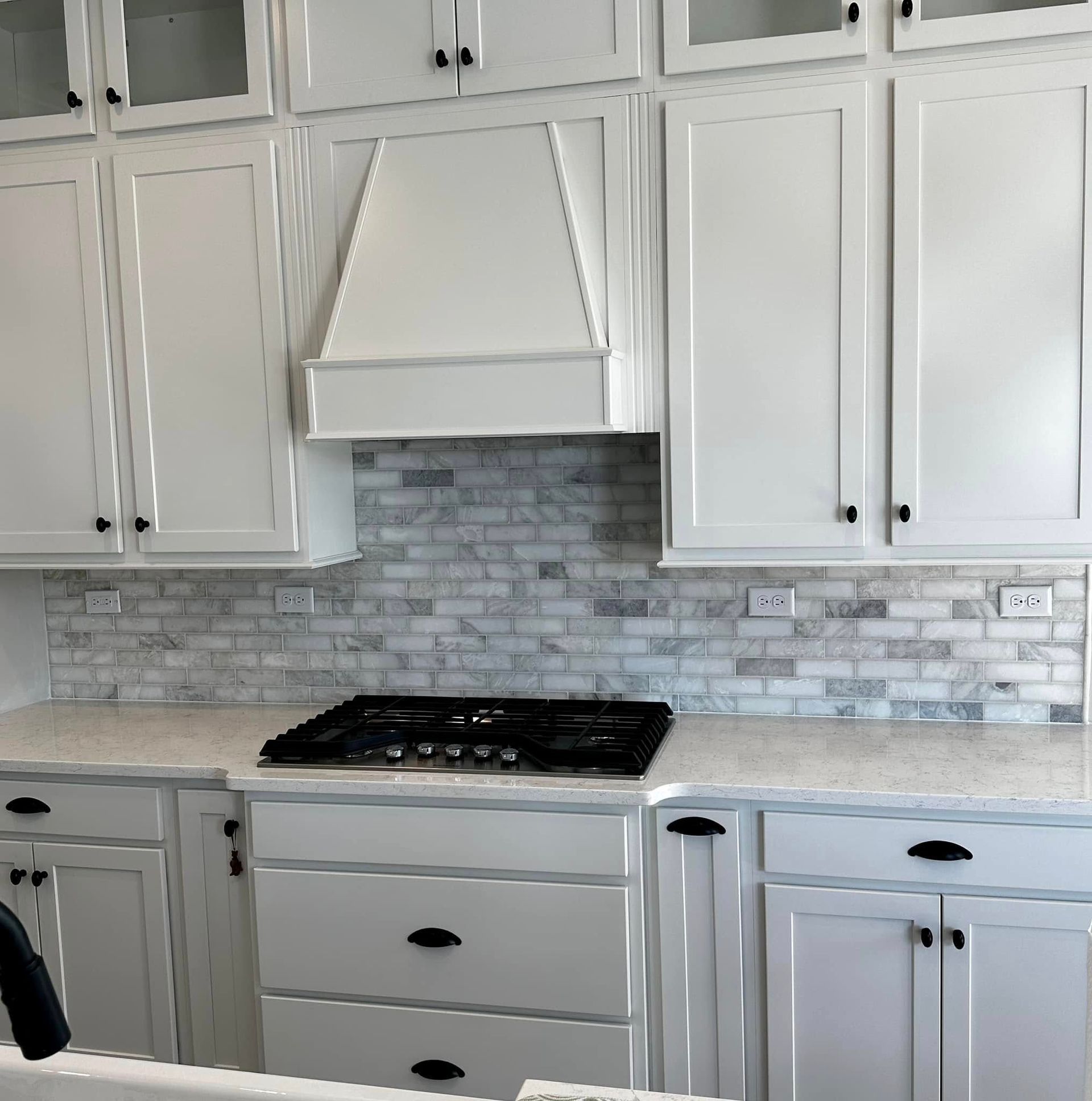 White kitchen cabinets with black hardware, white hood, and grey and white brick backsplash.