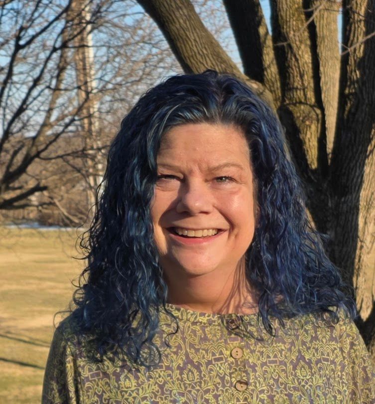 Smiling woman with blue hair standing outdoors by trees in sunlight