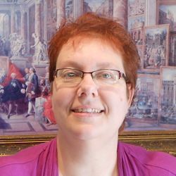 Person with glasses and short red hair smiling in front of a painted mural.