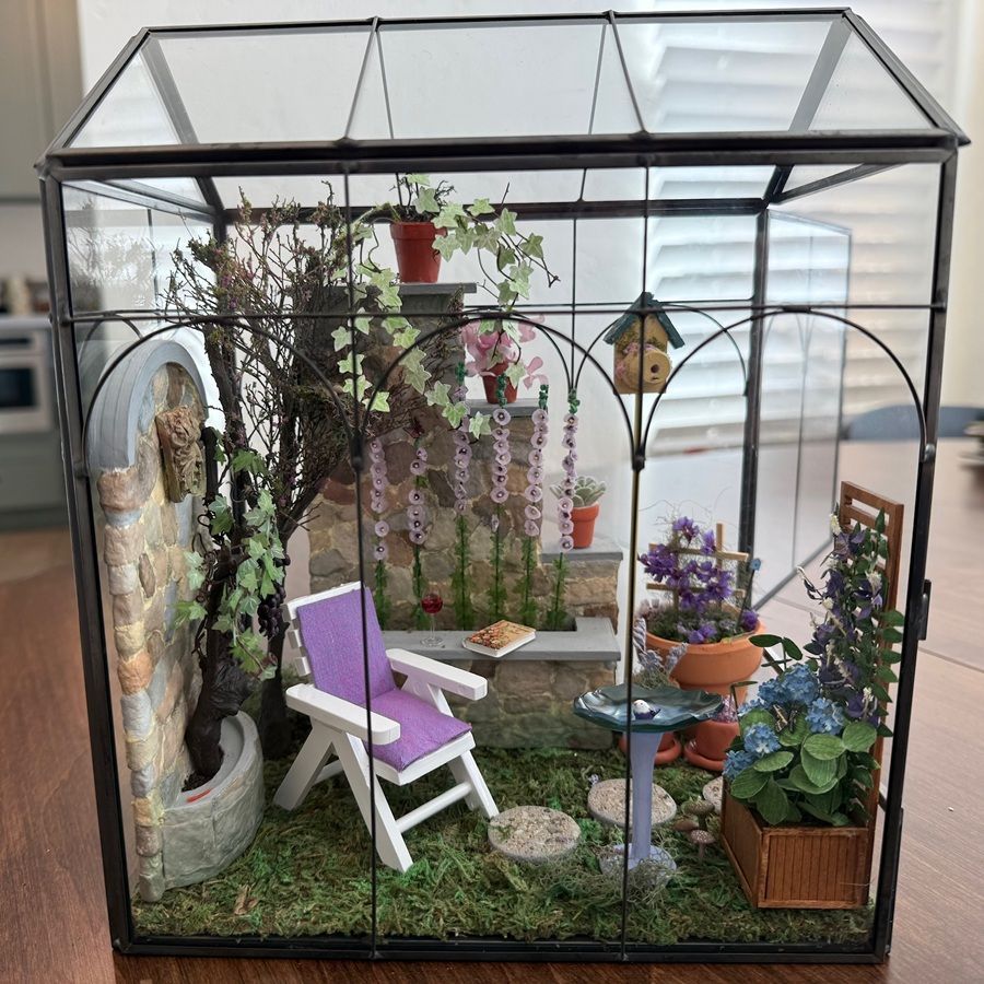 Miniature greenhouse garden with a white chair, purple accents, and potted flowers on grass inside.
