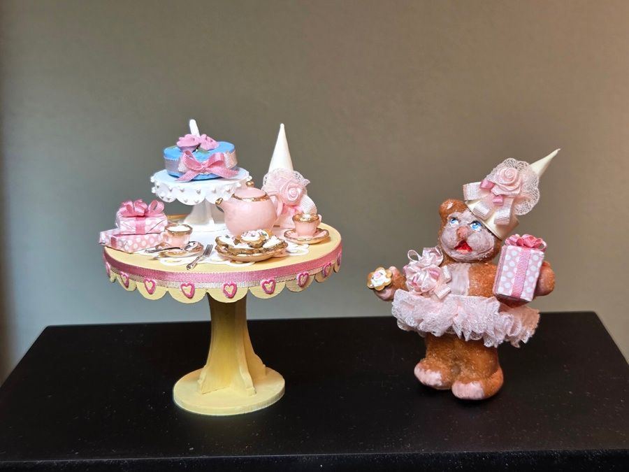 Two pink sugar figure displays on stands, one on a cake pedestal and one holding gifts, against a dark background