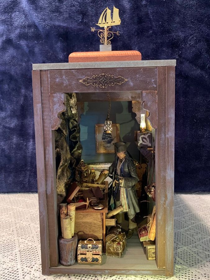 Miniature diorama in a wooden frame with a small ship finial, showing a figurine and stacked boxes inside.