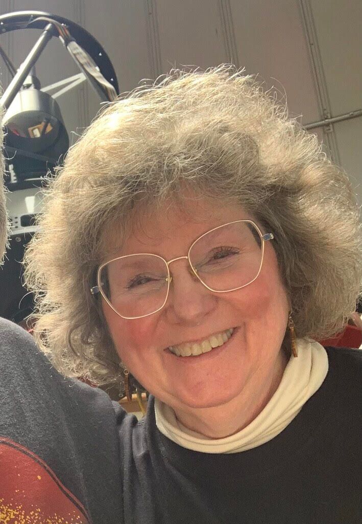 Smiling person with curly hair and glasses, wearing a black top and cream scarf in a casual indoor setting