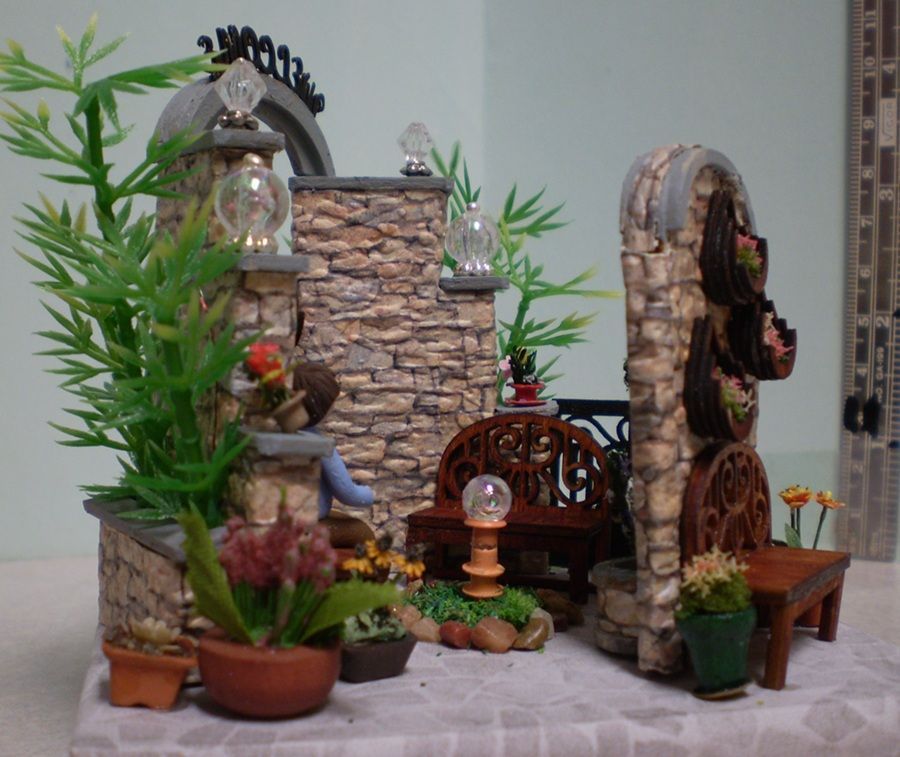 Miniature stone courtyard display with arches, plants, lanterns, and a small bench on a white cloth.