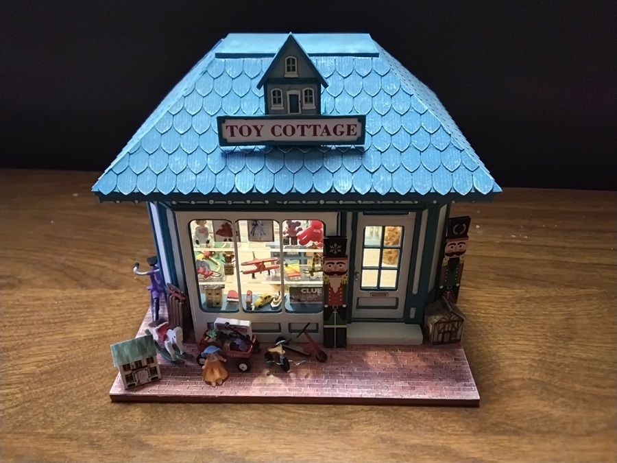 Miniature toy cottage with blue roof, lit storefront, and small figures on a wooden base.
