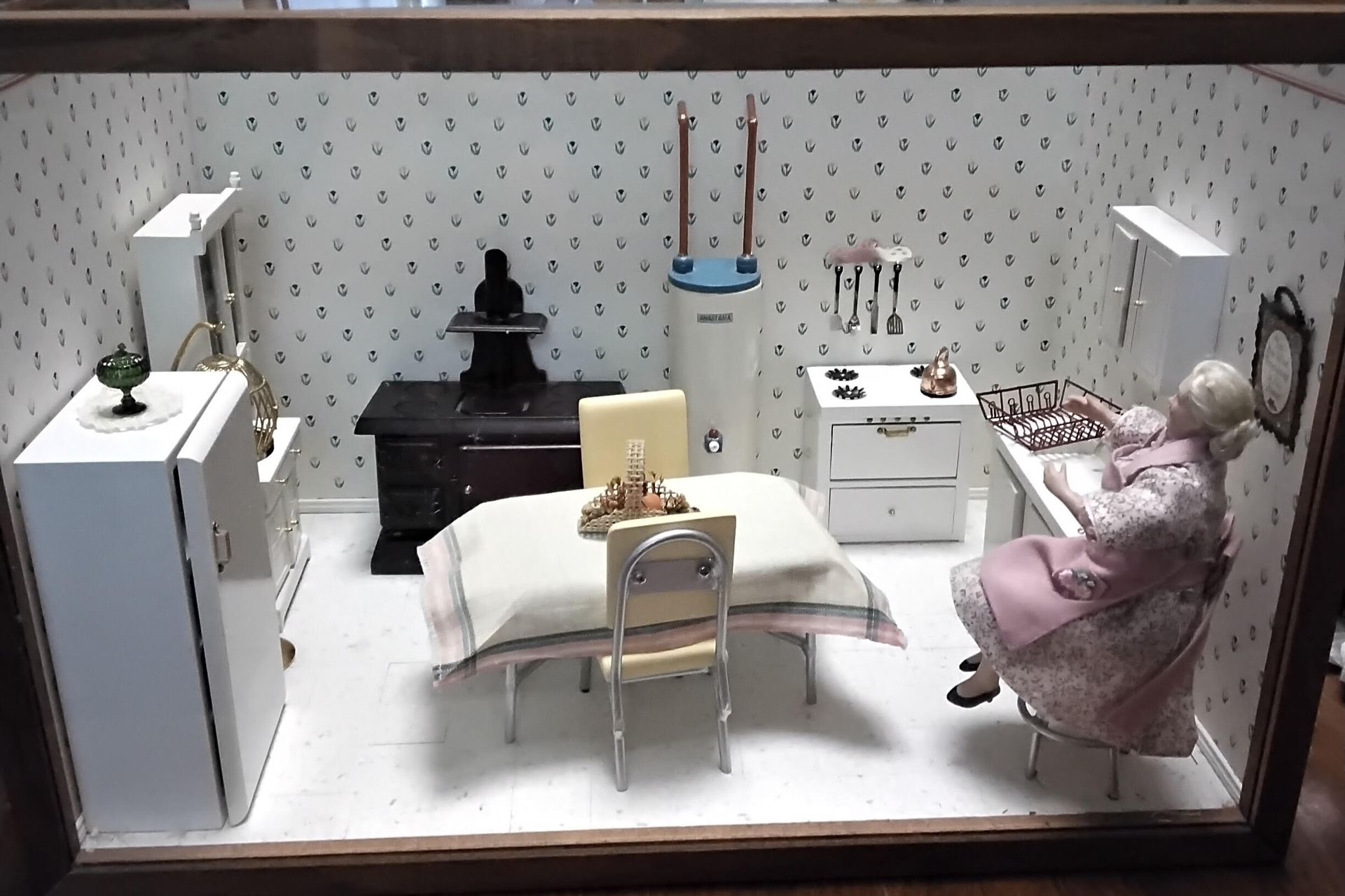 Miniature dollhouse dining room with table, chairs, white cabinets, and two dolls in pastel clothing