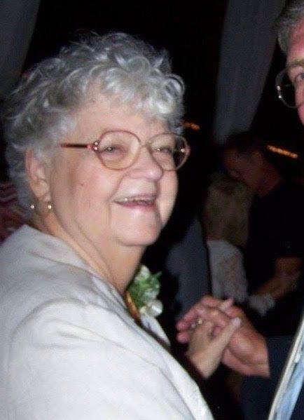 Smiling woman with curly gray hair and glasses, holding hands at a dimly lit event