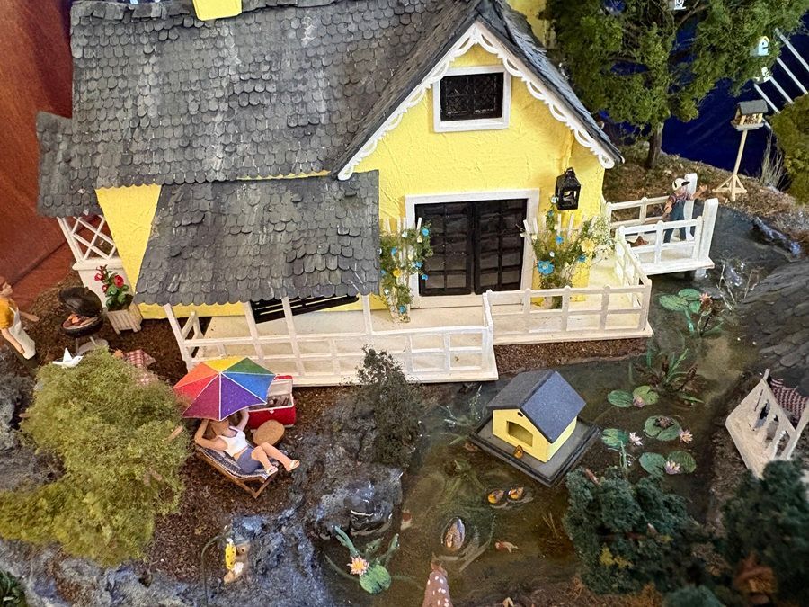 Miniature cottage with yellow walls, white porch, pond, and tiny people under a colorful umbrella