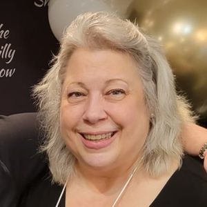 Smiling woman with gray hair in a black top, posing indoors with a dark backdrop and gold balloon behind her